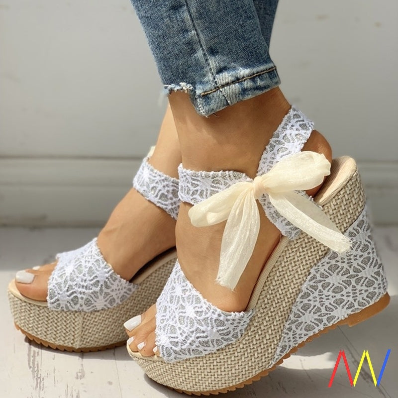 White lace wedge sandals with a bow worn with rolled-up jeans on a light background