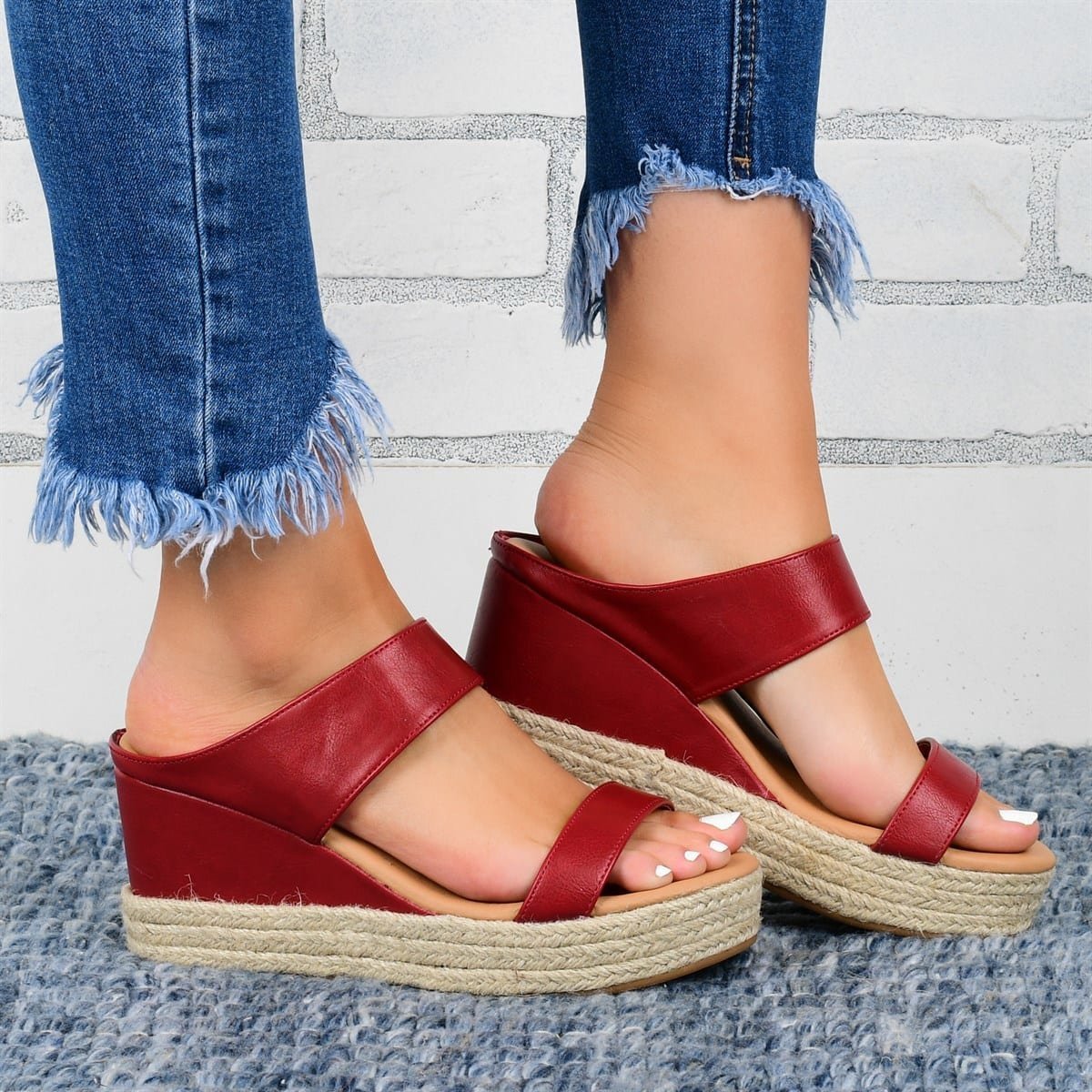 Red wedge sandals with jute sole worn with blue jeans against a light gray brick wall.