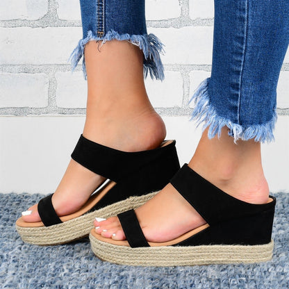 Black wedge sandals with jute sole worn with blue jeans against a light gray brick wall.