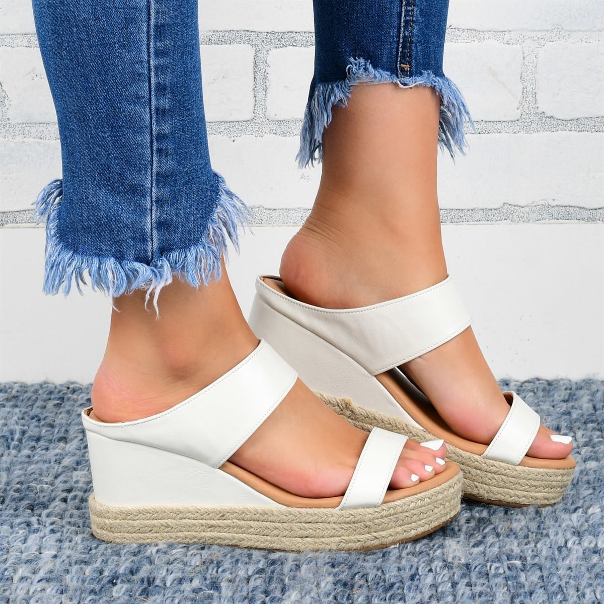 White wedge sandals worn with blue jeans against a light gray brick wall.