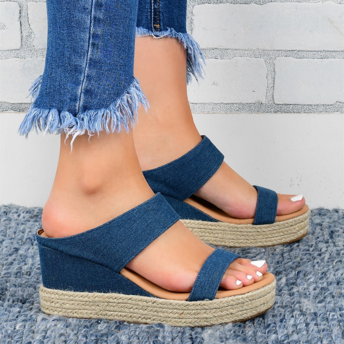Blue wedge sandals with jute sole worn with denim jeans against a gray brick wall.