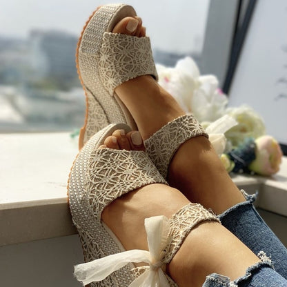 Beige wedge sandals with lace details worn by a person sitting by a window.