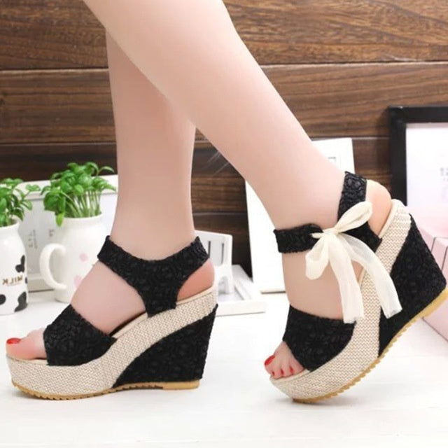 Black wedge sandals with white bows worn by a person on a blurred indoor background