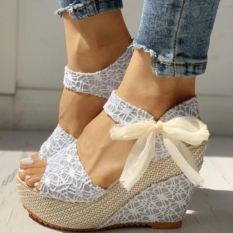 White lace wedge sandals with a bow worn by a person in denim jeans.