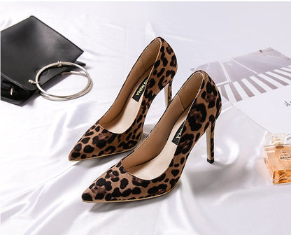 Leopard print high heels on a white surface with a black handbag and perfume bottle.
