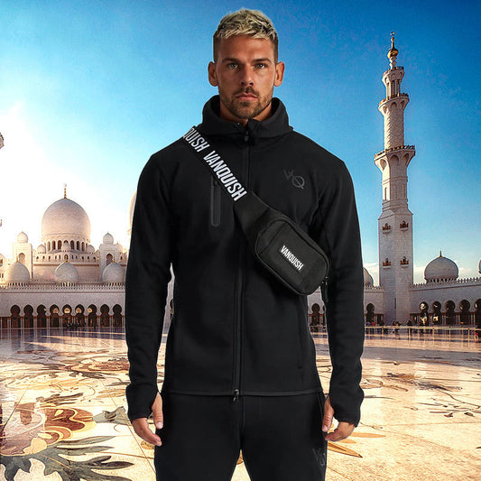 Man wearing a black jacket and carrying a black bag with 'Vandush' branding in front of a mosque.